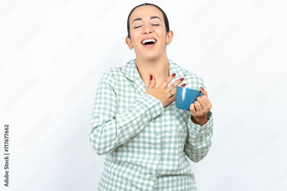 Beautiful young woman wearing green plaid pyjama and holding a cup expresses happiness, laughs pleasantly, keeps hands on heart Beautiful young woman wearing green plaid pyjama and holding a cup expresses happiness, laughs pleasantly, keeps hands on heart