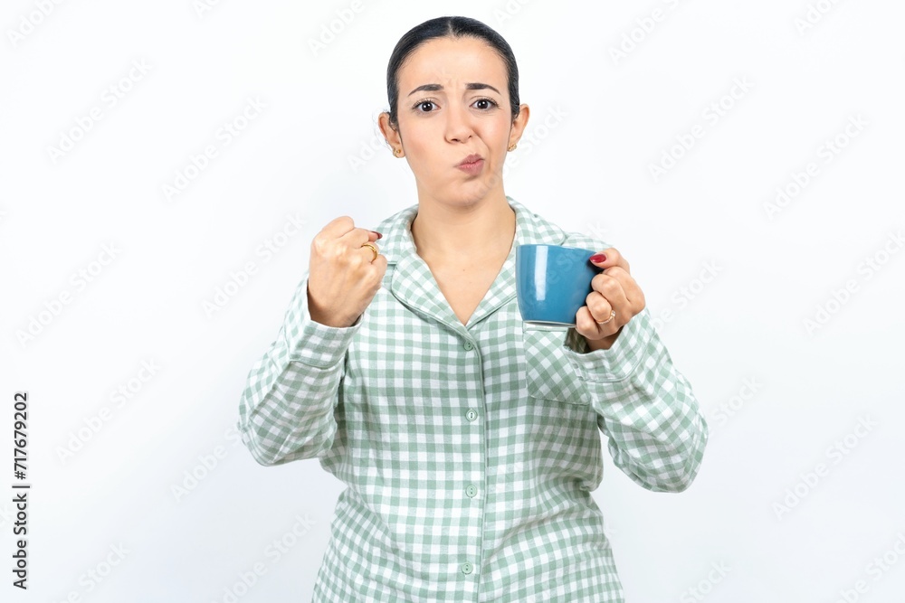 Displeased annoyed Beautiful young woman wearing green plaid pyjama and holding clenches fists, gestures pissed, ready to revenge, looks with aggression at camera stands full of hate, being pressured Displeased annoyed Beautiful young woman wearing green plaid pyjama and holding clenches fists, gestures pissed, ready to revenge, looks with aggression at camera stands full of hate, being pressured