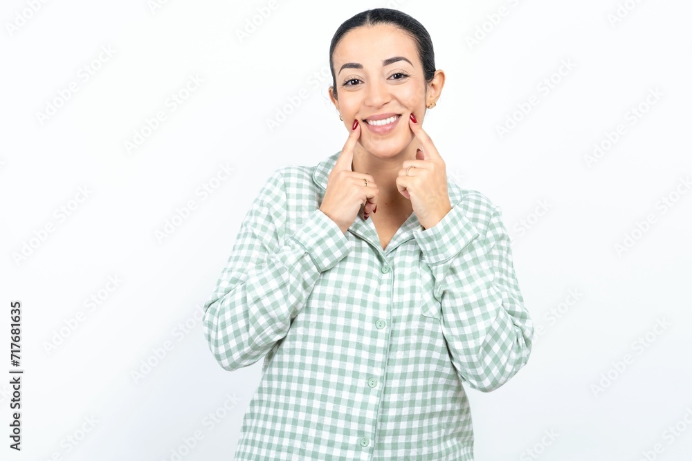 Happy Beautiful young woman wearing green plaid pyjama and holding a cup with toothy smile, keeps index fingers near mouth, fingers pointing and forcing cheerful smile Happy Beautiful young woman wearing green plaid pyjama and holding a cup with toothy smile, keeps index fingers near mouth, fingers pointing and forcing cheerful smile