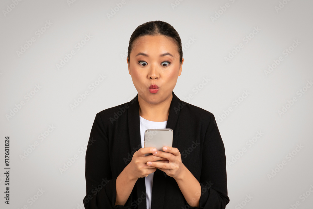 Surprised Asian businesswoman in a black blazer looking at her smartphone with an exaggerated expression of shock Surprised Asian businesswoman in a black blazer looking at her smartphone with an exaggerated expression of shock