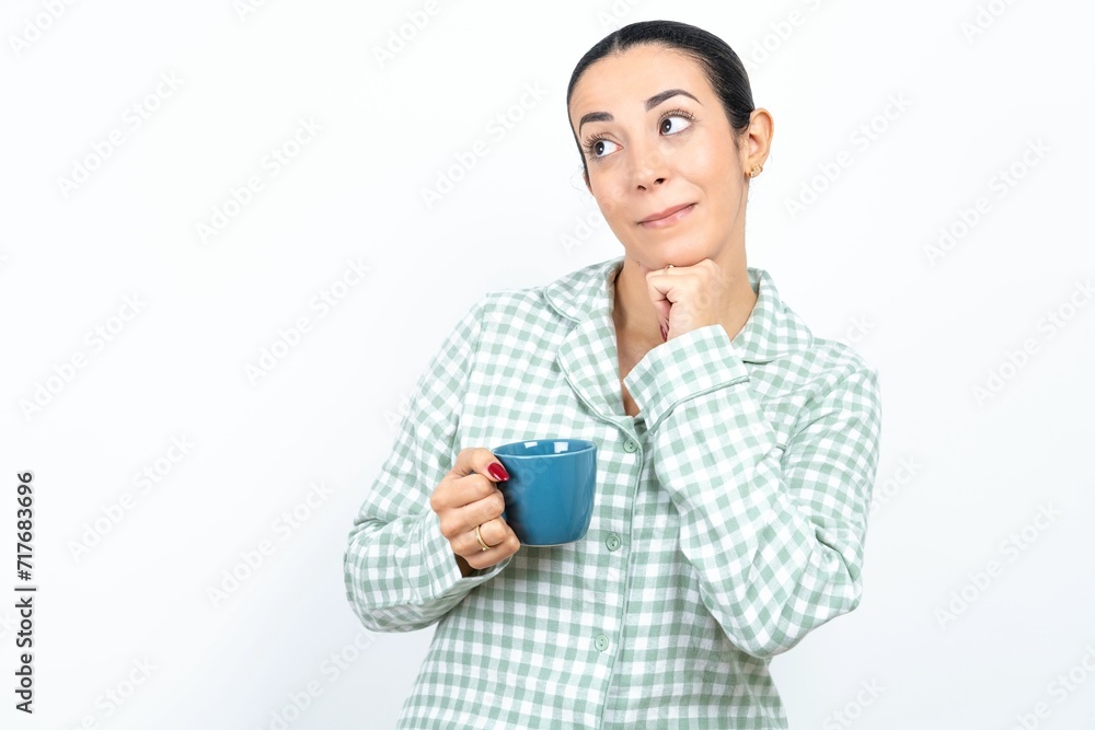 Curious Beautiful young woman wearing green plaid pyjama and holding a cup keeps hands under chin bites lips and looks with interest aside. Curious Beautiful young woman wearing green plaid pyjama and holding a cup keeps hands under chin bites lips and looks with interest aside.