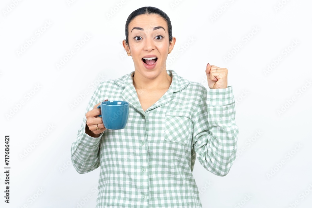 Beautiful young woman wearing green plaid pyjama and holding a cup points at empty space holding fist up, winner gesture. Beautiful young woman wearing green plaid pyjama and holding a cup points at empty space holding fist up, winner gesture.