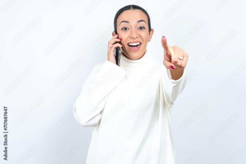 Positive Young beautiful woman wearing white turtleneck sweater indicates directly at camera has telephone conversation smiles broadly enjoys talking long hours. You join me Positive Young beautiful woman wearing white turtleneck sweater indicates directly at camera has telephone conversation smiles broadly enjoys talking long hours. You join me