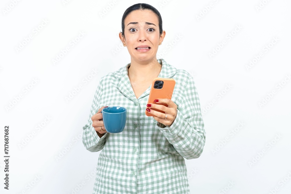 Afraid funny Beautiful young woman wearing green plaid pyjama and holding a cup holding telephone and bitting nails Afraid funny Beautiful young woman wearing green plaid pyjama and holding a cup holding telephone and bitting nails