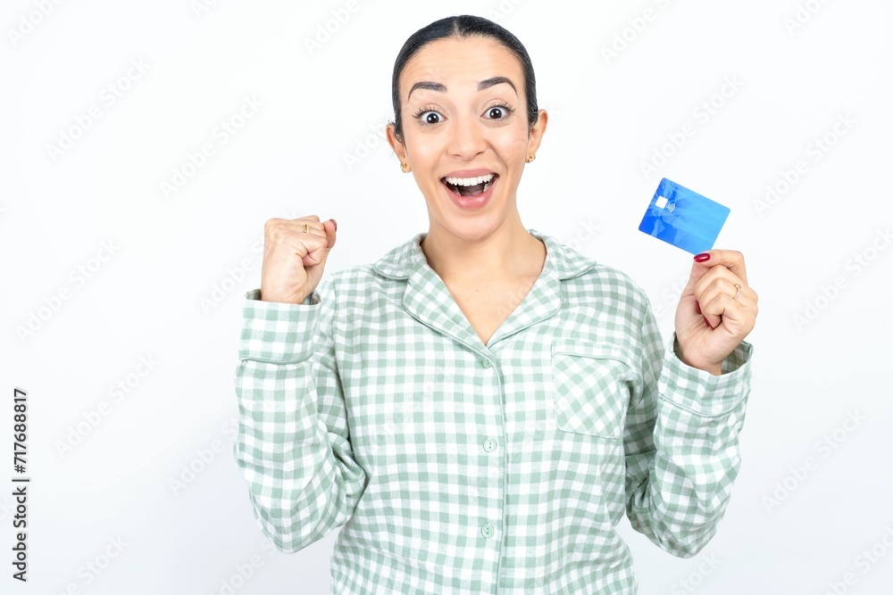 Photo of lucky impressed Beautiful young woman wearing green plaid pyjama and holding a cup arm fist holding credit card. Celebrated Photo of lucky impressed Beautiful young woman wearing green plaid pyjama and holding a cup arm fist holding credit card. Celebrated