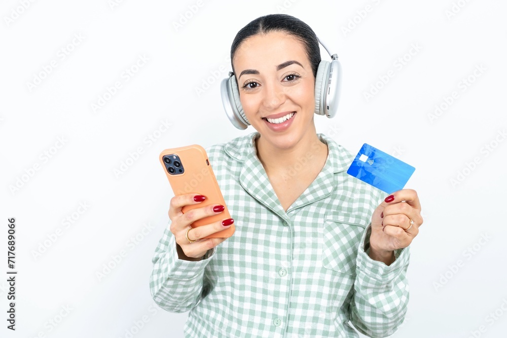 Beautiful young woman wearing green plaid pyjama and holding a cup wears stereo headphones on ears holds modern mobile phone and credit card Beautiful young woman wearing green plaid pyjama and holding a cup wears stereo headphones on ears holds modern mobile phone and credit card