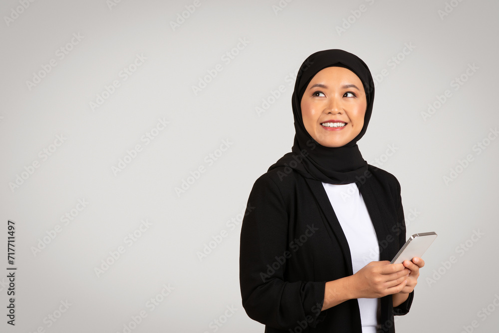 Thoughtful Muslim woman in a black hijab and blazer looks upwards, holding a smartphone Thoughtful Muslim woman in a black hijab and blazer looks upwards, holding a smartphone
