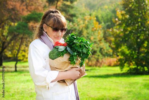 Stylish elderly caucasian brown-haired woman in sunglasses and white denim suit with craft paperr eco-friendly shopping bag with vegetables, herbs standing along an autumn street in sunny weather photo