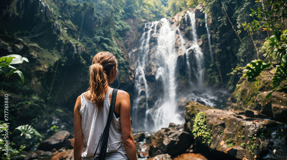 A woman with a backpack stands facing a majestic waterfall, surrounded by lush greenery in a tranquil forest setting.
A woman with a backpack stands facing a majestic waterfall, surrounded by lush greenery in a tranquil forest setting.