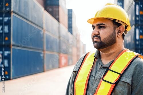 An attractive Indian male industrial engineer in yellow hard hat, safety vest with a blurred container yard in background. Working in the logistics center. Inspector, supervisor in container terminal.