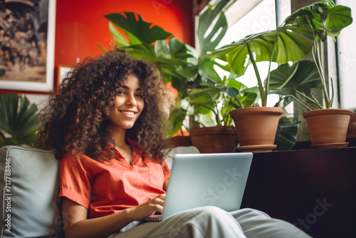 A joyful young latin american woman with curly hair comfortably uses a laptop.