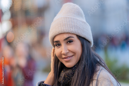 Close-up portrait of a cheerful woman wearing winter clothes, city lights softly out of focus. © JoseIMartin