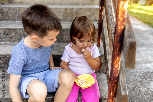 Two kids, aged 6 and 2, eating popcorn together on front of their house. Young kids eating popcorn outdoors, on a sunny day. Close-up.