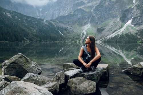 Beautiful young brunette woman in stylish outfit sitting on the rocks, stones on the shore and admiring beautiful view of green hills and mountains on Morskie Oko lake, High Tatras, Zakopane, Poland photo