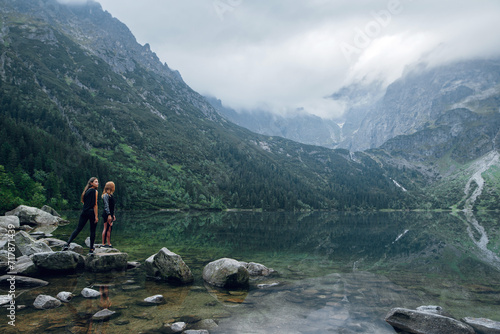 Two women, friends standing on the stony shore of lake Moskie Oko with scenic view of mountains with clouds and fog. Rysy mountains, Tatras. Poland, Slovakia. Rear view. Travel and hiking concept