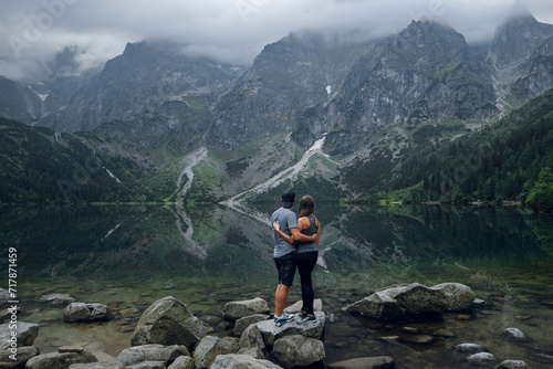 Young couple of man and woman standing on the stony shore of lake Moskie Oko with scenic view of mountains with clouds and fog. Rysy mountains, Tatras. Poland, Slovakia. Rear view.