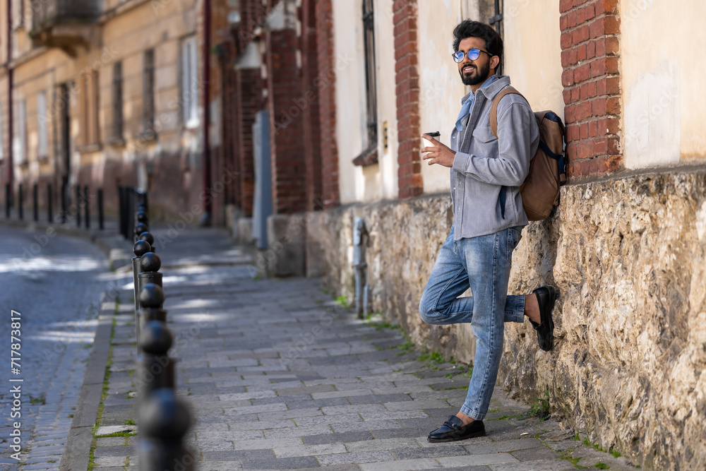 Young handsome hindu man standing near the wall with a coffee cup in hand