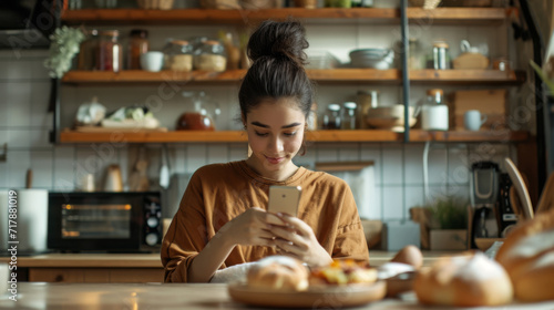 young woman with a high puff hairstyle is smiling down at her smartphone, seated at a table with a sandwich on a plate in front of her