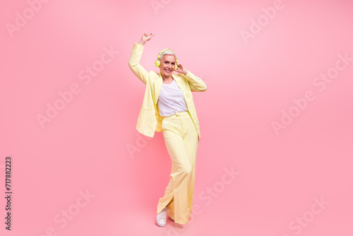 Full size photo of cheerful satisfied woman dressed yellow suit listen music in headphones dancing isolated on pink color background