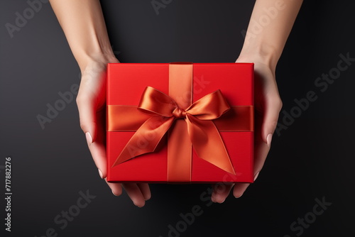 Top view of hands holding craft red paper gift box with ribbon bow on isolated black background. Generated AI. © jirayut