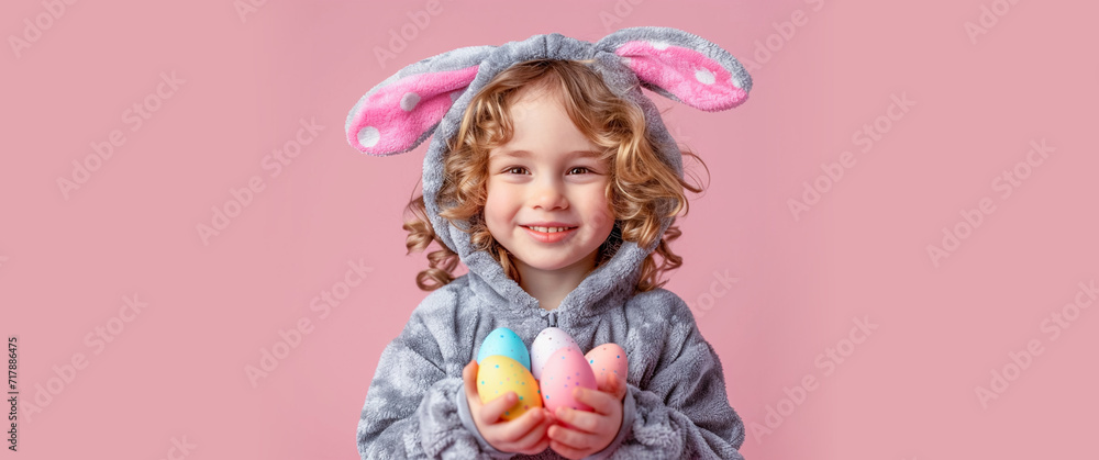 A cute little smiling blond curly-haired boy in a rabbit costume with ears with Easter colored colored eggs in his hands on a pink background. Banner, copy space A cute little smiling blond curly-haired boy in a rabbit costume with ears with Easter colored colored eggs in his hands on a pink background. Banner, copy space