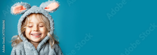 A happy blond little boy with a sweet smile and closed eyes in gray Easter bunny pajamas with fluffy ears on a blue background. Banner, a place to copy