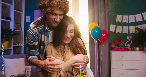 Photography Caucasian young attractive wife sitting at the table and her husband bringing birthday cake with candles which she blowing