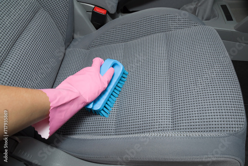 Woman hand in rubber protective glove holding brush and cleaning gray textile car seat. Care about auto interior. Point of view shot. photo