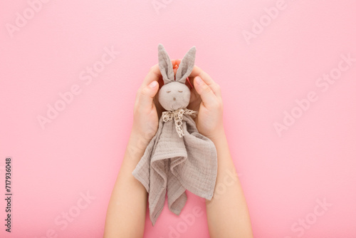 Little girl hands holding light gray cloth bunny toy on light pink table background. Pastel color. Point of view shot. Top down view.