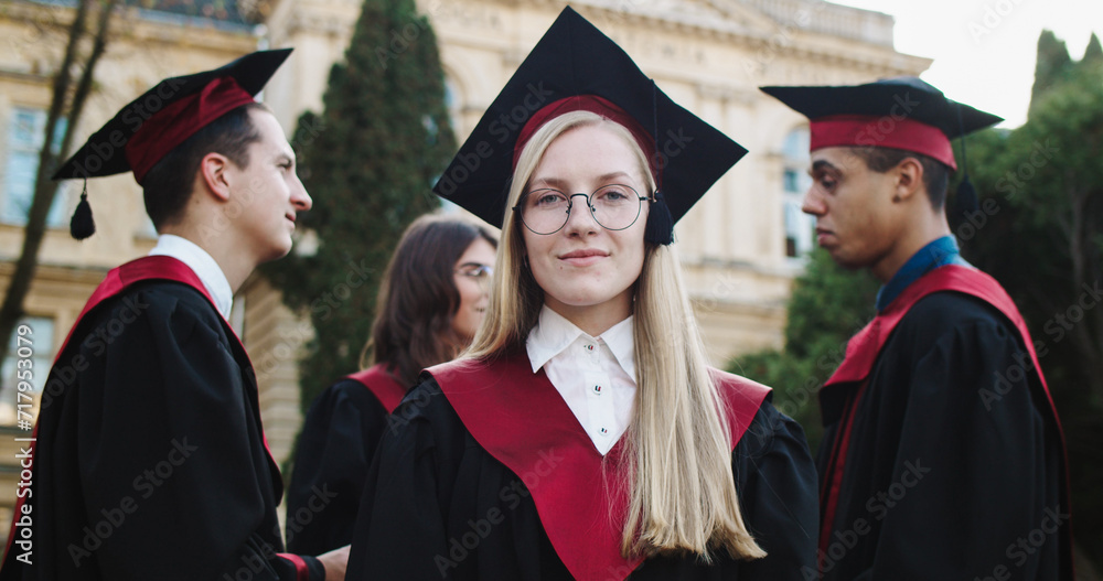 Portrait of the young pretty Caucasian mlond student in glasses, academic cap and gown smiling to the camera at the university building during graduation ceremony. Outside. Close up.