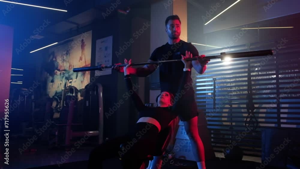 A female athlete is engaged in an incline bench press workout at the gym with her trainer, illuminated by a dramatic fusion of blue and red lights with mist enhancing the intense atmosphere A female athlete is engaged in an incline bench press workout at the gym with her trainer, illuminated by a dramatic fusion of blue and red lights with mist enhancing the intense atmosphere