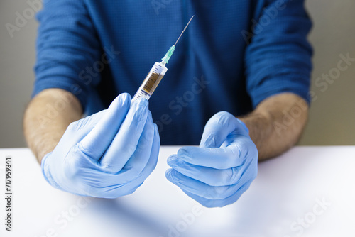 The hands of the doctor or nurse, on a white table and wearing blue gloves, hold a syringe with a needle with injectable medication inside. Horizontal.
