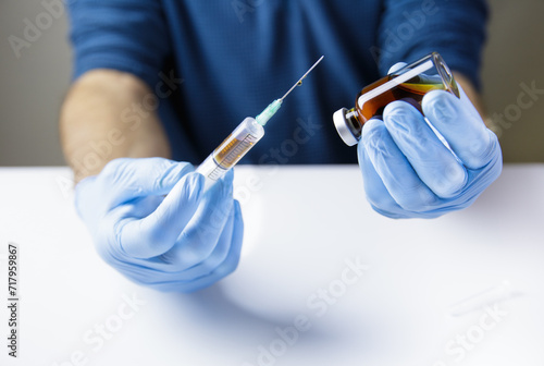 Canvas Print The hands of the doctor or nurse, on a white table and wearing blue gloves, hold a syringe with a needle and the bottle of injectable medicine