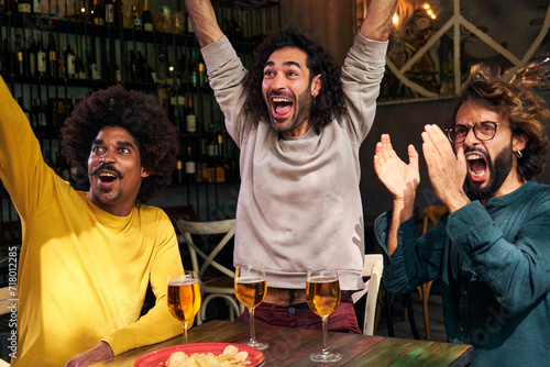 Multiethnic group of three male friends watching soccer match at a bar. Celebrating happily a score with beers and chips, gesturing arms raised and screaming goal looking at tv screen.