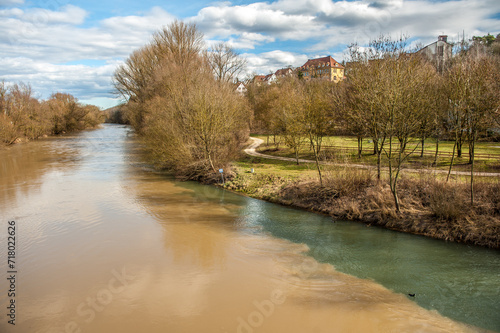 Schmaler Fluss mit klarem Wasser fliesst in breiten Fluss mit braunem Wasser vor Landschaft mit Fussweg kahlen Bäumen und Gebäude