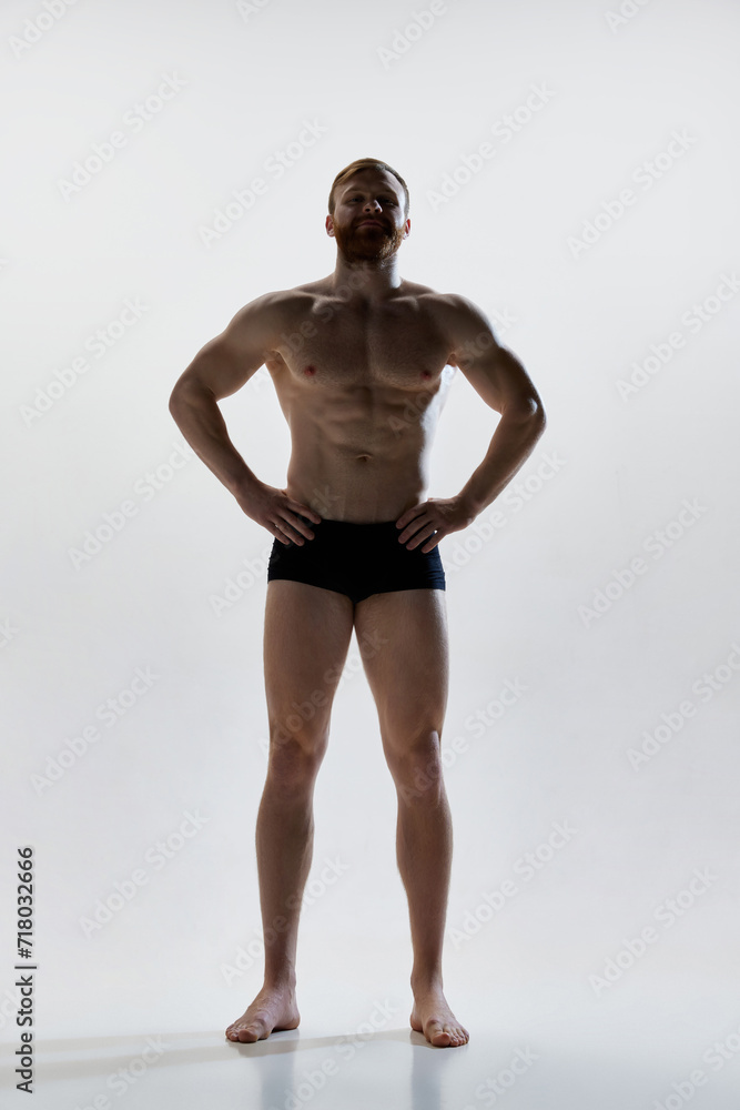 Fototapeta premium Full length portrait of young attractive man with perfect body curves posing in underwear against white background. Concept of natural beauty, aesthetic of body, male health, masculinity.