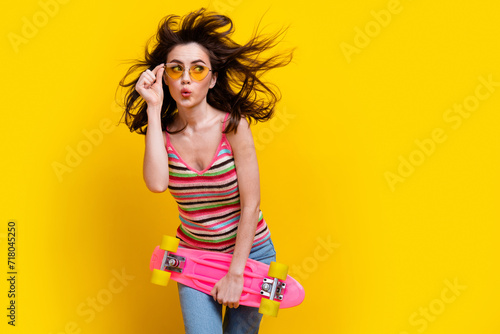 Portrait of excited girl fluttering hair touch glasses hold skateboard look at offer empty space isolated on yellow color background