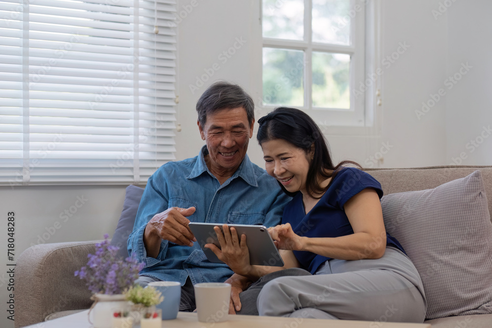 Cheerful Asian couple using network video call tablet sitting together on sofa at home Asian elderly husband sitting with elderly wife shopping online or video calling on tablet, relaxing. Cheerful Asian couple using network video call tablet sitting together on sofa at home Asian elderly husband sitting with elderly wife shopping online or video calling on tablet, relaxing.
