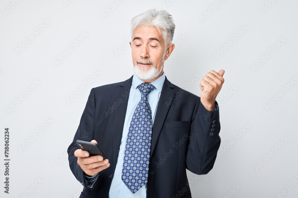 Portrait of elderly businessman in suit with mobile phone in hand celebrates victory, winning money. Person with smartphone isolated on white background. Portrait of elderly businessman in suit with mobile phone in hand celebrates victory, winning money. Person with smartphone isolated on white background.