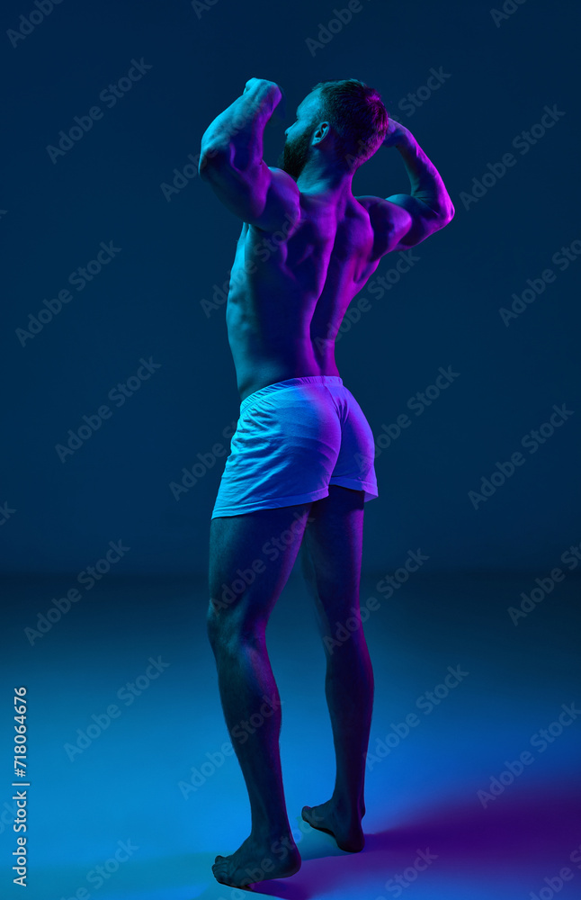 Rear view photo of young, fit naked man demonstrated his perfect back shape in neon light against studio background. Concept of beauty and fashion, art, aesthetic of body, masculinity.
