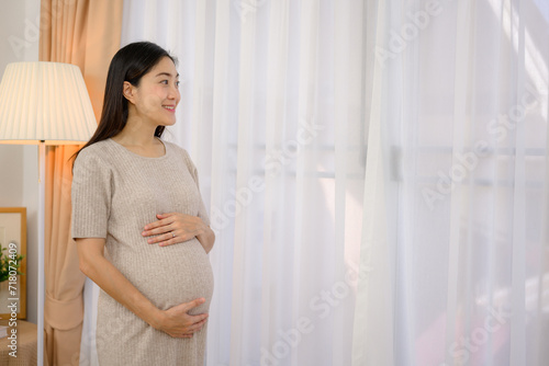 A beautiful pregnant Asian woman is smiling and happy at home carrying her large belly while standing by the white window in her home.