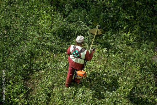 The gardener in overall and safety helmet trims overgrown grass by grass cutter