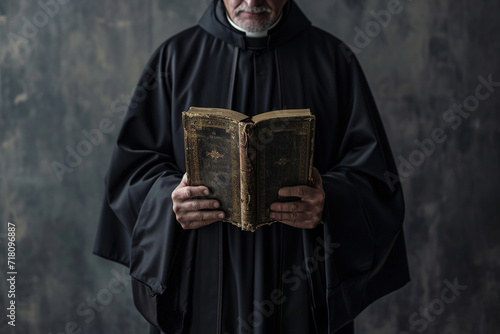 a catholic christian church priest wearing black cassock robe holding the holy bible book in his hands. face seen. isolated on dark grey / black background. photo