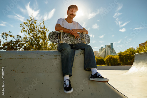Smiling young male skateboarder holding skateboard sitting on ramp in skate park  photo
