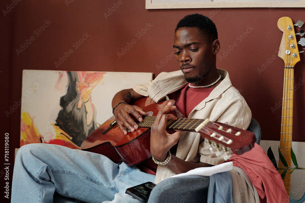 African American guy in casualwear sitting in armchair in front of camera and playing acoustic guitar against wall with abstract paintings