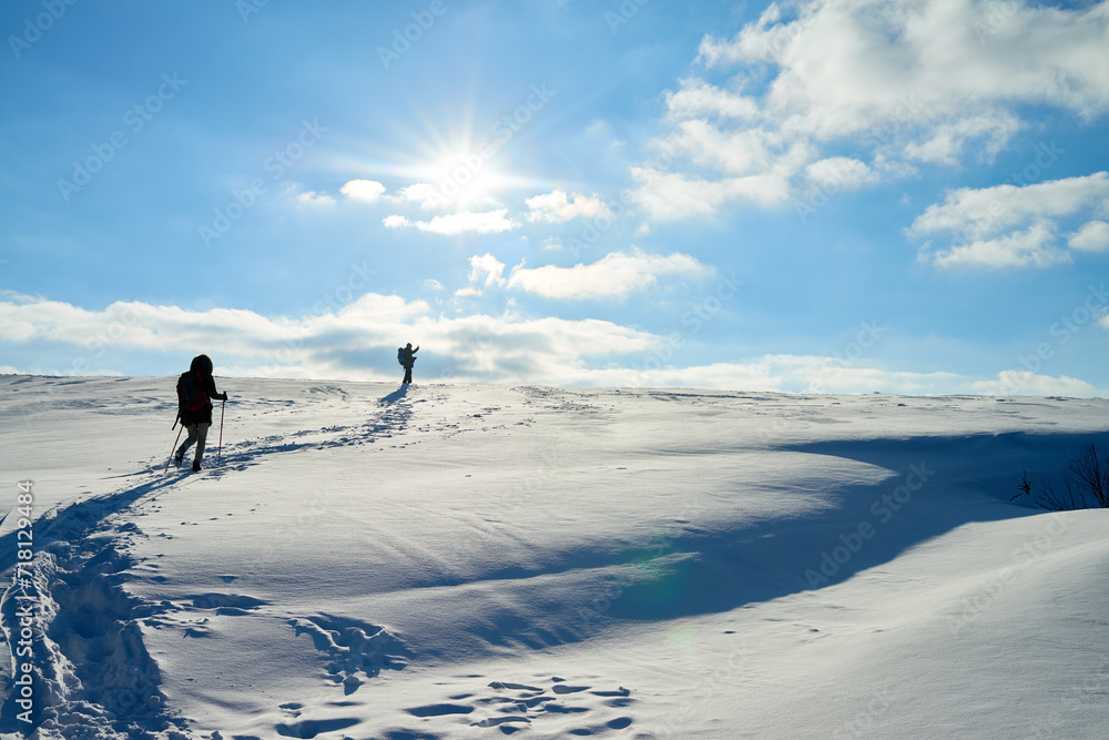Unrecognizable travelers using trekking poles while climbing to mountains on snow-covered slope under shining sun