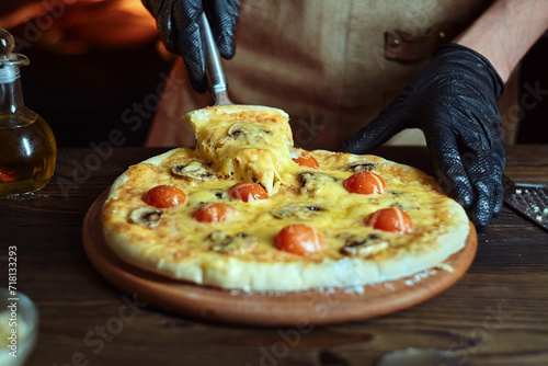 A guy in black gloves picks up a pizza triangle. Pizza cooked in the oven with mushrooms and tomatoes.