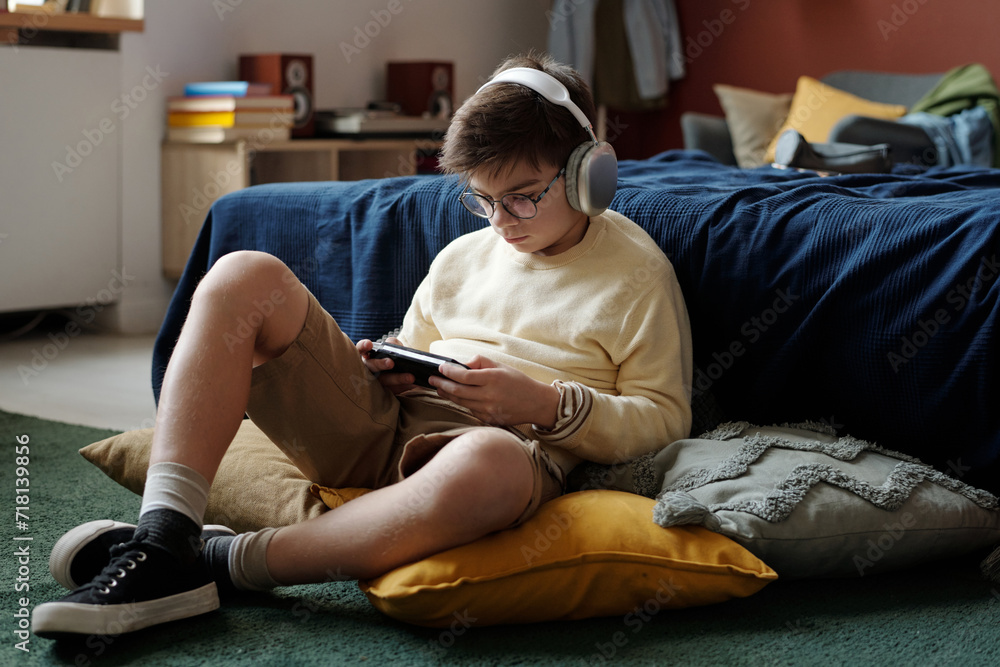 Obraz premium Modern schoolboy in casualwear sitting on the floor by bed and playing video game while looking at screen of mobile phone in gamepad