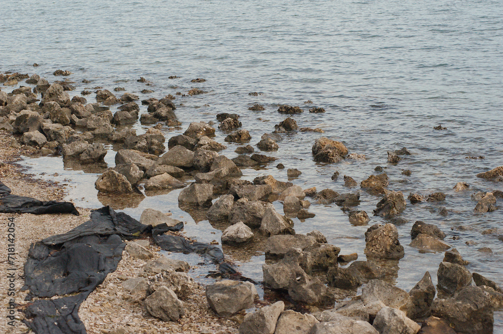 View over multi colored rocks with long narrow black cloth trash washed ashore tangled in the rocks on shore. Water in background with small rippled waves. Late afternoon on a cloudy day. View over multi colored rocks with long narrow black cloth trash washed ashore tangled in the rocks on shore. Water in background with small rippled waves. Late afternoon on a cloudy day.