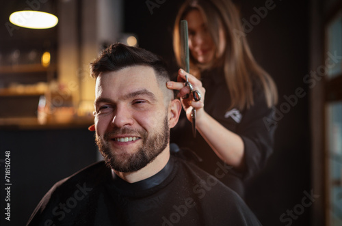 Smiling bearded Man portrait while undercut hairstyle hairdressing in Hair Salon by young hairdresser female. Modern low light black style barber shop interior. Haircare service small business concept photo